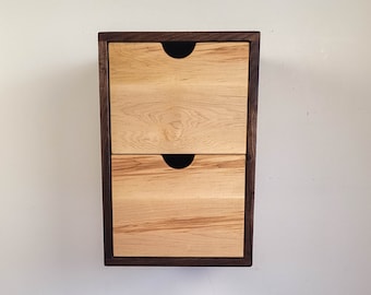 Puede incluir: Un armario de pared de dos cajones con marco de madera oscura y cajones de madera clara. Los cajones tienen tiradores negros.