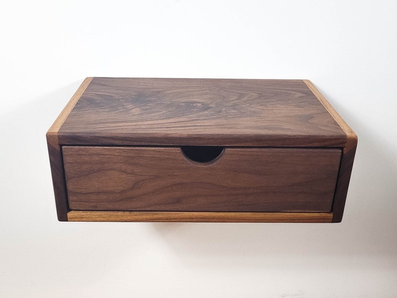 May include: A dark brown wooden floating shelf with a single drawer. The shelf has a light brown wood trim around the edges.