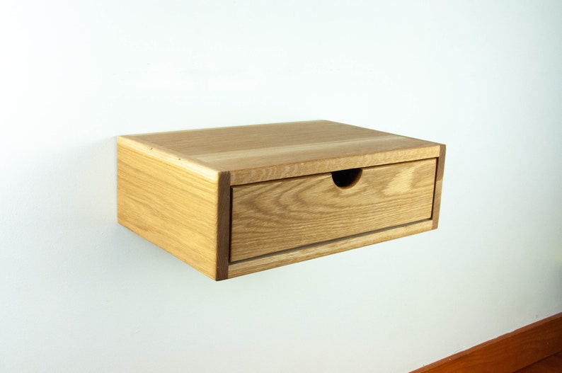 May include: A light wood floating bedside table with a single drawer. The drawer has a slightly rounded cutout for the handle.
