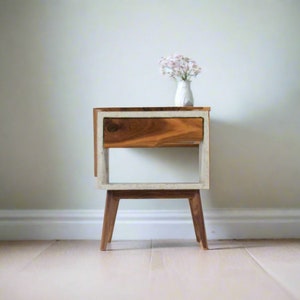 May include: A wooden nightstand with a white painted frame and a single drawer. The nightstand has four legs and is sitting on a light wood floor.