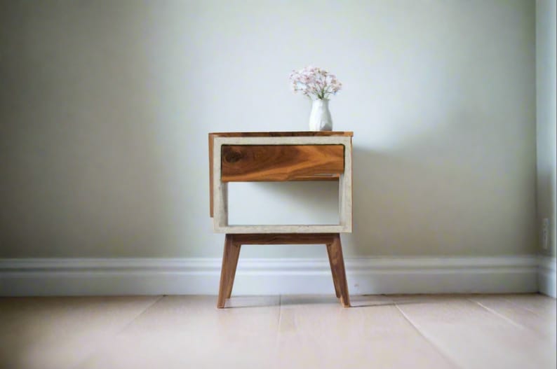 May include: A wooden nightstand with a white painted frame and a single drawer. The nightstand has four legs and is sitting on a light wood floor.