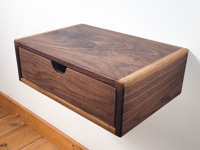 May include: A dark brown wooden floating bedside table with a single drawer. The drawer has a simple, rectangular cutout for a handle.
