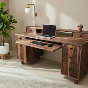 May include: A dark wood desk with multiple drawers, a pull-out keyboard tray, and a hutch. A laptop, keyboard, and mouse sit on the desk. A small potted plant is on the hutch. A large potted plant is to the left.