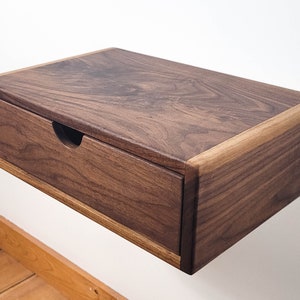 May include: A dark brown wooden floating bedside table with a single drawer. The bedside table has a light brown wood trim around the edges.