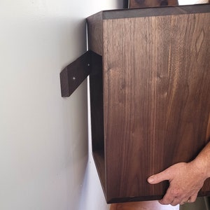May include: A dark brown wooden wall shelf with a simple design. The shelf is being installed on a white wall.