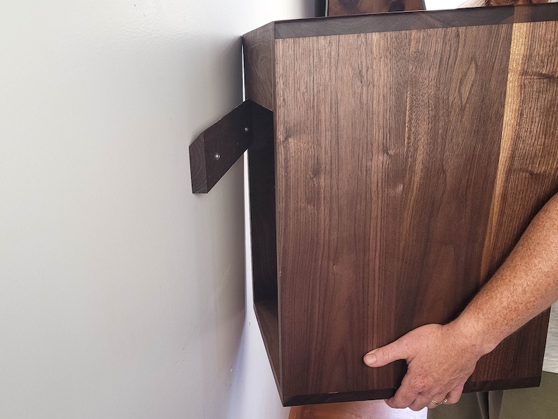 May include: A dark brown wooden wall shelf with a simple design. The shelf is being installed on a white wall.