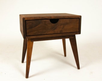 May include: A dark brown wooden bedside table with a single drawer and four tapered legs. The drawer has a black handle.
