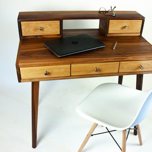 May include: A wooden desk with four drawers and a shelf. The desk has a dark brown finish and is made of walnut wood. A laptop computer and a pen are on the desk. A white chair with wooden legs is in the foreground.