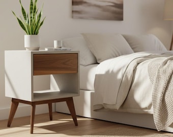 May include: A bedside table featuring a white frame, a single wooden drawer, and an open storage shelf underneath. The table is supported by four tapered wooden legs and holds a white potted snake plant and a small white cup on its top surface.