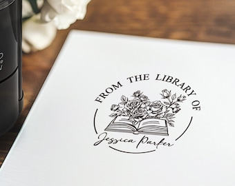 May include: A black COLOP printer R 40N stamp next to a white paper with a floral design and the text "FROM THE LIBRARY OF" and a name. The stamp is on a wooden surface with white flowers in the background.