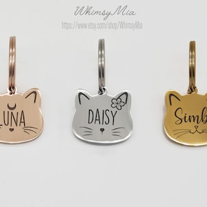 May include: Three cat-shaped pet tags in rose gold, silver, and gold. Each tag features a cat face design with engraved names: "Luna", "Daisy", and "Simba". The tags have a loop for attaching to a collar.