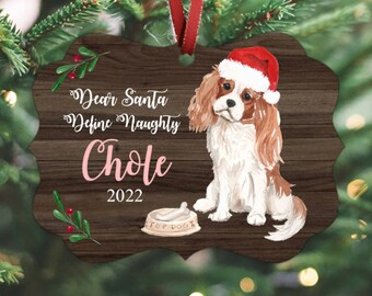 May include: A wooden Christmas ornament with a watercolor illustration of a Cavalier King Charles Spaniel wearing a Santa hat. The ornament has the text "Dear Santa Define Naughty Chole 2022" and a dog bowl with a bone.