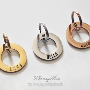 May include: Three personalised pet ID tags. One is gold-coloured with the name "LUNA", another is silver with "BELLA", and the third is rose gold with "WILLOW" and a paw print. Each tag has a ring for attachment.
