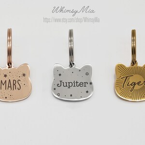 Op de afbeelding: Drie metalen huisdierlabels in de vorm van een kattenkop. E&eacute;n is ros&eacute;goud met het woord "Mars", &eacute;&eacute;n is zilver met "Jupiter" en &eacute;&eacute;n is goud met "Tiger". Elk label heeft een ring. De winkelnaam "Whimsy Mia" staat bovenaan.