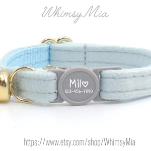 May include: A light blue cat collar with a gold bell and a gray clasp. The clasp has the name "Mil" and a phone number. The collar is made of a soft material and has a light blue interior.