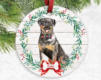 Op de afbeelding: Ronde kerstversiering met een Rottweiler-hond met een bril en een geruite vlinderdas, tegen een witte achtergrond met houtnerf. De hond wordt omringd door een groene krans met rode bessen. Een rood lint en de naam "Dawn" zijn ook aanwezig.