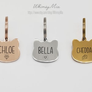Op de afbeelding: Drie katvormige huisdier-ID-tags in ros&eacute;goud, zilver en goud. Elke tag heeft een gegraveerde naam: "CHLOE" met een diamant, "BELLA" met een hart en "CHEDDAR" met een kaasicoon. De tags hebben een ring voor bevestiging.