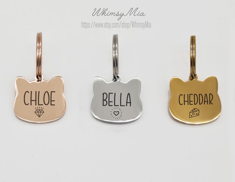 Op de afbeelding: Drie katvormige huisdier-ID-tags in ros&eacute;goud, zilver en goud. Elke tag heeft een gegraveerde naam: "CHLOE" met een diamant, "BELLA" met een hart en "CHEDDAR" met een kaasicoon. De tags hebben een ring voor bevestiging.