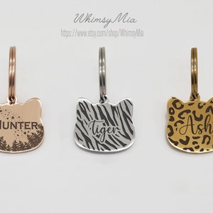 Op de afbeelding: Drie metalen dierenpenningen in de vorm van een kattenkop. E&eacute;n is ros&eacute;goud met "HUNTER" en een bosontwerp. De tweede is zilver met een zebraprint en "Tiger". De derde is goudkleurig met een luipaardprint en "Ash".