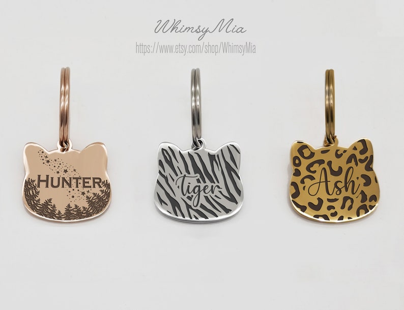 Op de afbeelding: Drie metalen dierenpenningen in de vorm van een kattenkop. E&eacute;n is ros&eacute;goud met "HUNTER" en een bosontwerp. De tweede is zilver met een zebraprint en "Tiger". De derde is goudkleurig met een luipaardprint en "Ash".