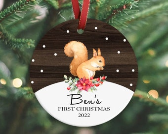 Op de afbeelding: Ronde kerstversiering met een aquarel eekhoorn ontwerp. De versiering heeft een donkere houtnerf achtergrond met witte sneeuwvlokken. De onderste helft is wit met de tekst "Ben's FIRST CHRISTMAS 2022". Een rood lint is bevestigd.