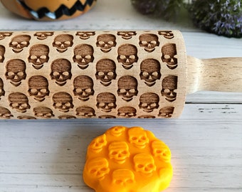 Op de afbeelding: Een houten deegroller met een herhalend schedelpatroon. De roller is lichtbruin met een handvat van natuurlijk hout. Een oranje, schedelvormig object staat op de voorgrond. Het thema is Halloween en bakken.