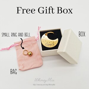 May include: A gold-colored crescent moon pendant with star details and the word "COPPER" in a white gift box. A small ring with a bell is in a pink bag. The text "Free Gift Box" is at the top.