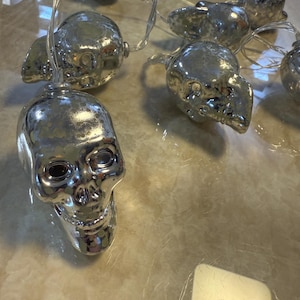 Puede incluir: Una cadena de luces plateadas con forma de calaveras. Cada calavera tiene detalles faciales y está ensartada en un cable transparente. Las luces son reflectantes y parecen ser de metal, perfectas para la decoración de Halloween.