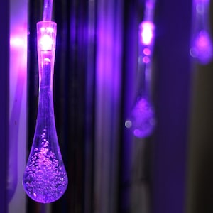 May include: Purple string lights with teardrop-shaped bulbs. The bulbs are filled with small, clear beads that reflect the light.