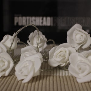 May include: White artificial flower string lights with a warm white glow. The flowers are arranged in a cluster and are connected by a clear wire. The lights are perfect for adding a touch of elegance to any space.