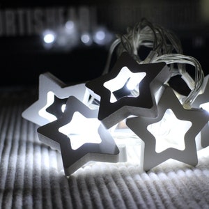 Puede incluir: Una cadena de luces con forma de estrella blanca. Las estrellas de madera emiten un brillo cálido. Las luces están conectadas por un fino cable blanco y descansan sobre una superficie blanca texturizada.