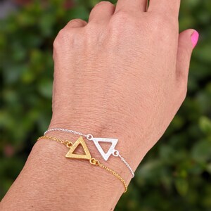Puede incluir: Dos delicados brazaletes de triángulo, uno dorado y otro plateado, en la muñeca de una persona. Los brazaletes son simples y elegantes, perfectos para el uso diario.
