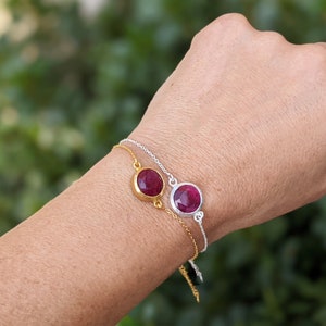 May include: Two delicate bracelets with a red gemstone charm on each. The bracelets are made of silver chain and have a gold accent on one of the charms.