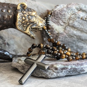 May include: A black cross pendant necklace with a tiger's eye and hematite beaded chain. The necklace is displayed on a rock with a bronze animal sculpture.