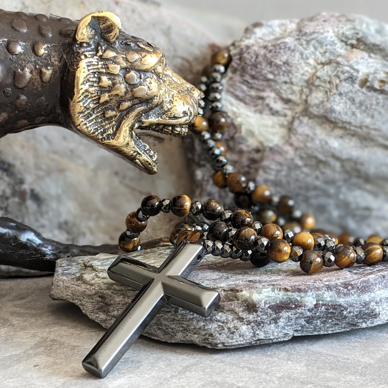 May include: A black cross pendant necklace with a tiger's eye and hematite beaded chain. The necklace is displayed on a rock with a bronze animal sculpture.