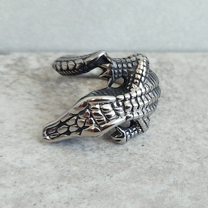 May include: A silver ring featuring a detailed alligator design. The alligator is depicted in a curled position with its head and tail visible.