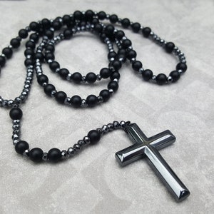 May include: A black beaded necklace with a large, black cross pendant. The necklace is made of two strands of beads, one with round beads and one with faceted beads.