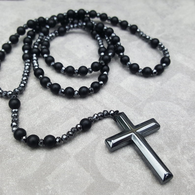 May include: A black beaded necklace with a large, black cross pendant. The necklace is made of two strands of beads, one with round beads and one with faceted beads.