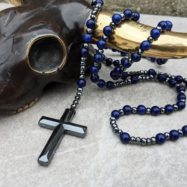 May include: A black cross pendant hangs from a beaded necklace with dark blue and black beads. The necklace is draped over a brown, textured surface.