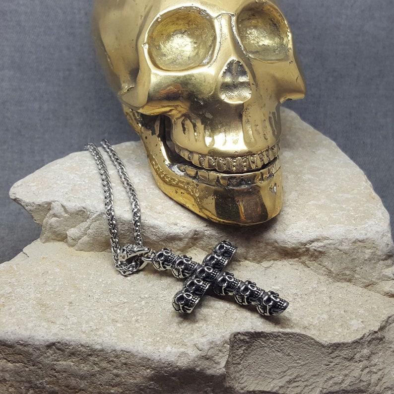 May include: A silver chain necklace with a skull-shaped cross pendant. The cross is made of multiple skulls, each with a detailed design. The necklace is displayed on a rock next to a gold skull.