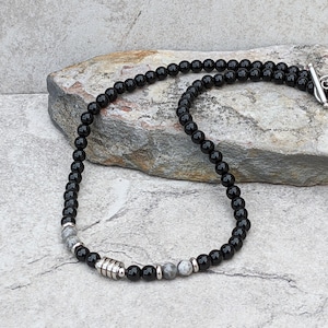 May include: A black beaded necklace with silver accents. The necklace is made of round black beads and silver spacer beads. The necklace is lying on a grey rock.