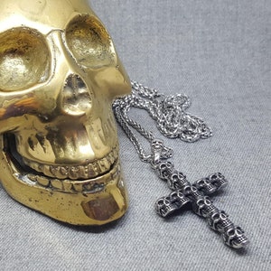 May include: A silver skull-shaped pendant necklace with a silver chain. The pendant is a cross with multiple skulls on it. The necklace is lying on a gold skull.