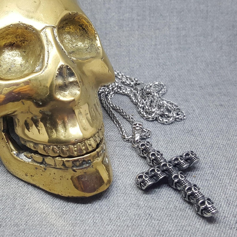 May include: A silver skull-shaped pendant necklace with a silver chain. The pendant is a cross with multiple skulls on it. The necklace is lying on a gold skull.
