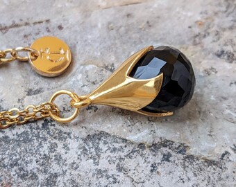 Puede incluir: Un collar de cadena de oro con un colgante de piedra negra. El colgante es una piedra negra facetada engastada en un engaste de oro que se asemeja a una flor.