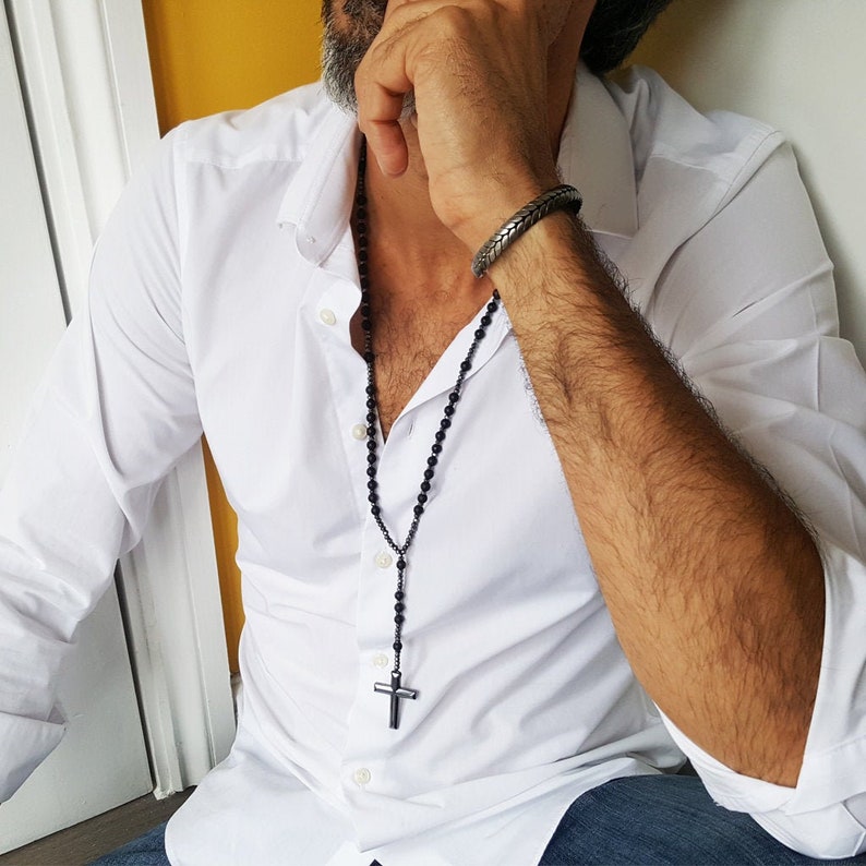 May include: A black beaded necklace with a black cross pendant. The necklace is worn by a person wearing a white button-down shirt.