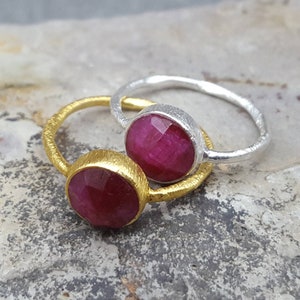 以下が含まれることがあります： 赤い宝石がセットされた銀の指輪が2つ。左側の指輪は金メッキされています。指輪は、質感のある灰色の表面の上に置かれています。