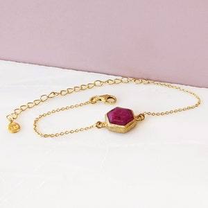 May include: A gold chain bracelet with a single, hexagonal, dark pink gemstone.