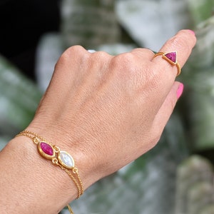 Könnte beinhalten: Ein goldenes Kettenarmband mit zwei ovalen Edelsteinen, einem roten und einem weißen. Das Armband wird am Handgelenk einer Person getragen. Ein goldener Ring mit einem dreieckigen roten Edelstein wird an derselben Hand getragen.