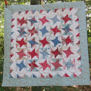 May include: A handmade quilt featuring a star pattern in red, blue, and floral fabrics on a white background. The quilt has a light blue border with a floral design and is hanging outdoors.