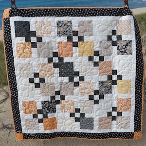 May include: A handmade quilt with a patchwork design. The quilt features squares in various patterns and colors, including black, orange, tan, and cream. The squares are arranged in a grid pattern and connected by black squares. The quilt has a black and white patterned border.